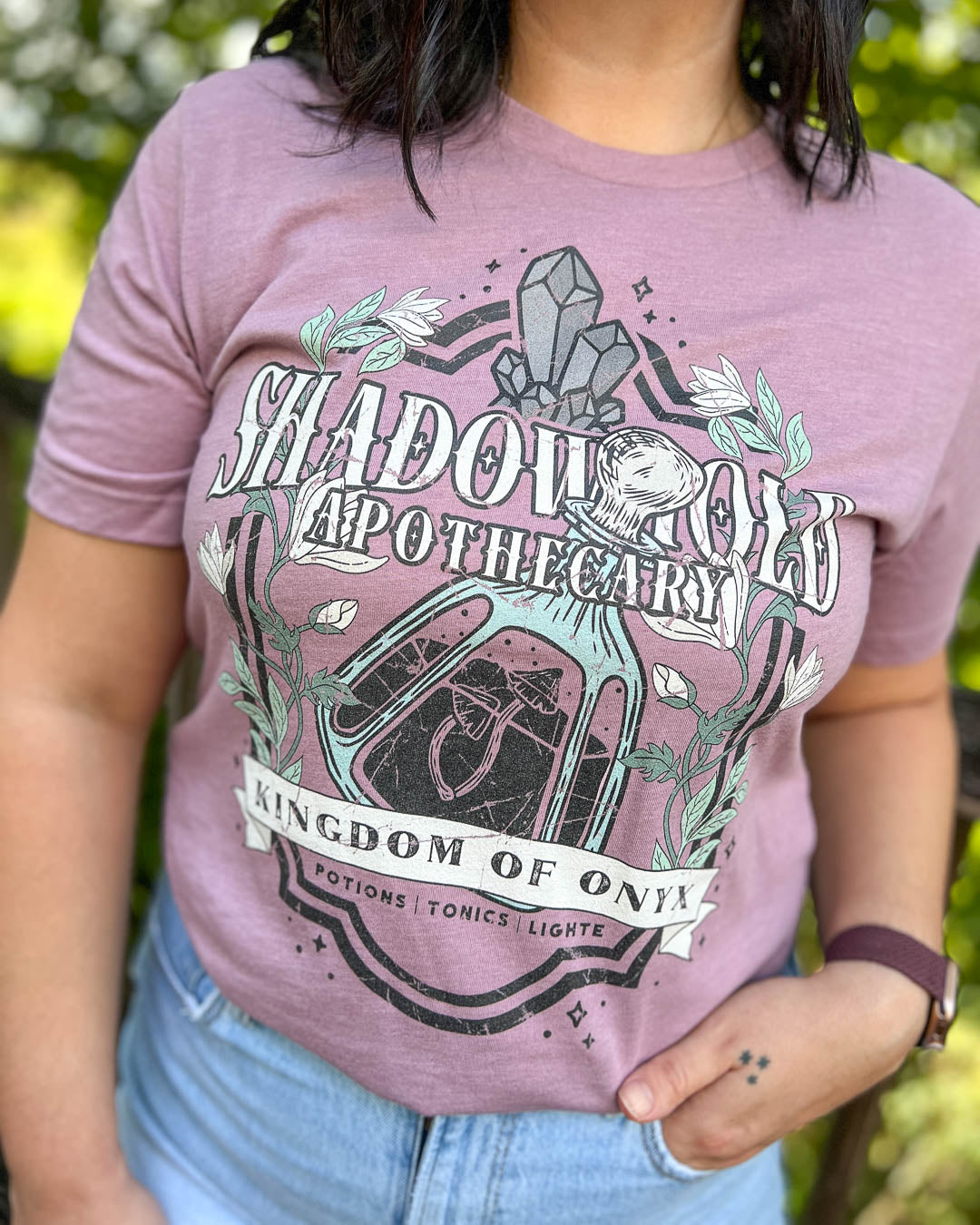 Kate Golden shirt featuring a Shadowhold Apothecary logo designed with a glass bottle full of a potion, gems, stars, and flowers for A Dawn of Onyx fans.