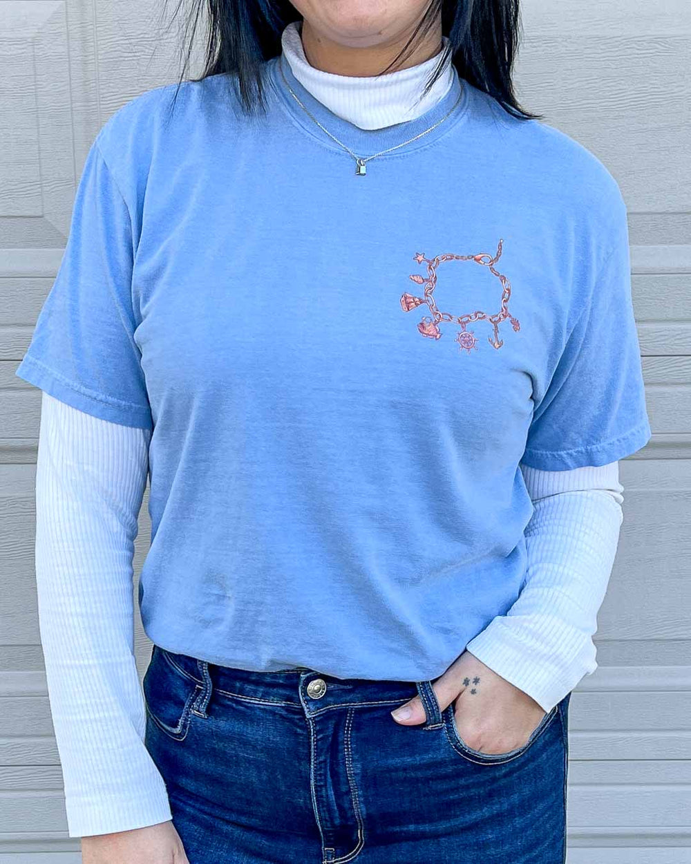 Woman wearing the blue Siren's Lure shirt, featuring a bronze charm bracelet with nautical charms on the top left chest, over a white long-sleeve.