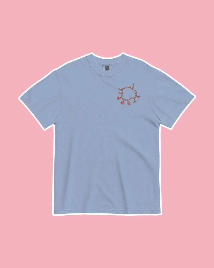 Blue t-shirt on pink background with an illustrated charm bracelet printed on the top left chest inspired by Half City by Kate Golden.