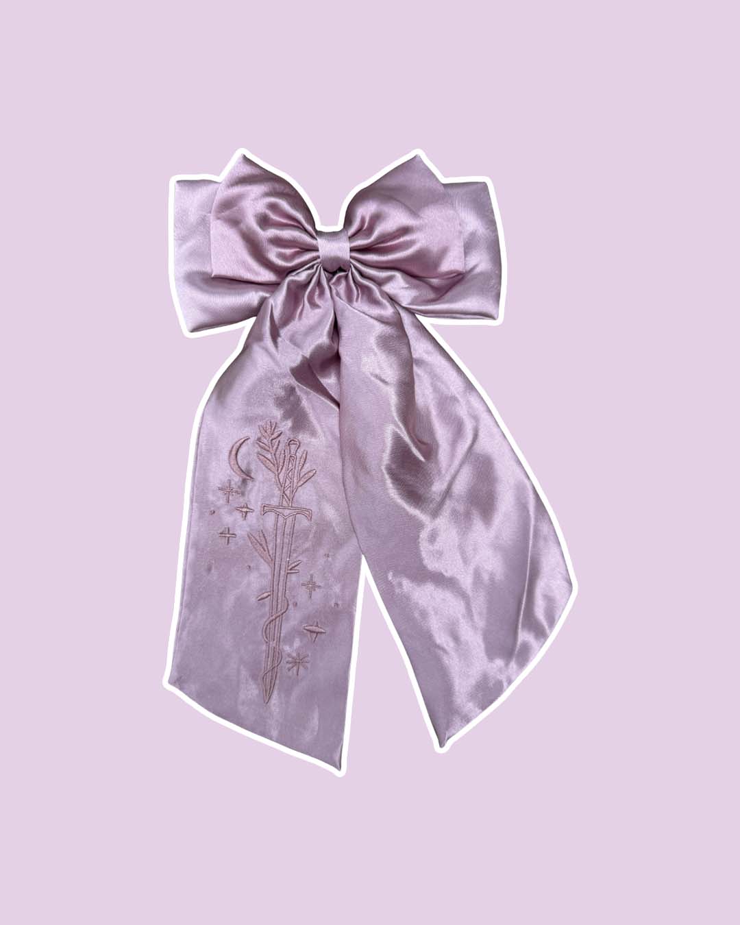 Purple satin hair bow clip with an embroidered design of a sword wrapped in vines surrounded by the moon and stars on the left tail on a purple background.