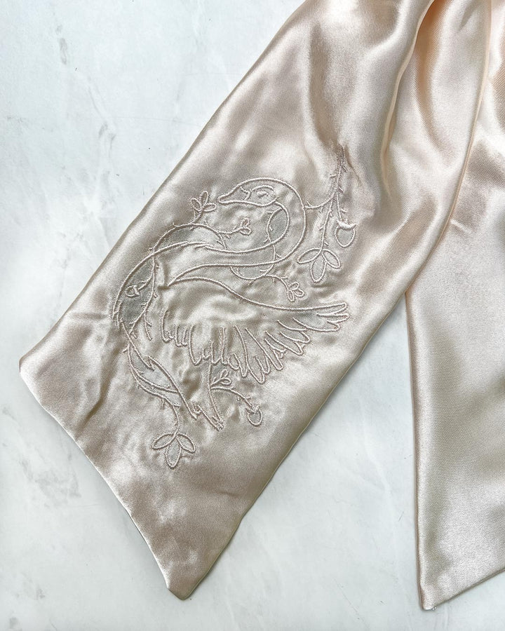 Close up of the embroidered swan wrapped in ivy on the a bookish satin hair bow in a shiny ivory color.