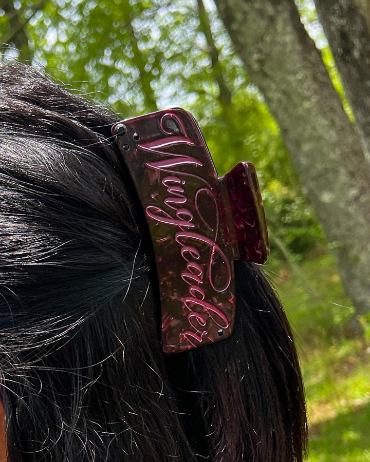 A maroon pearlescent bookish hair clip with pink script saying &