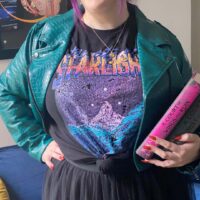 person standing wear the City of Starlight Vintage Location Tee from A Court of Thorns and Roses Inside Green Jacket with a skirt while holding the books in one hand and on their hip with the other
