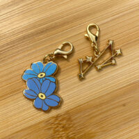 Two charms on a wood background. One charm is two blue colored flowers. The other charm is 7 gold stars connected by lines.
