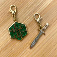 Image of two gold charms, one in a hexagonal shape with the text "she elf" and the other is in the shape of a sword. Both are inspired by Lord of the Rings.