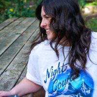 person sitting at a picnic table wearing leggings and night court inspired boardwalk airbrush tee in the woods