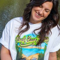 person sitting next to a lake wearing the north bear shores beach read boardwalk airbrush inspired white boyfriend tee
