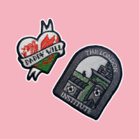 Embroidered patch shaped like a heart with a Welsh dragon with the text "daddy will" on a denim jacket. Embroidered patch shaped with the text "the london institute" and gray building on pink background
