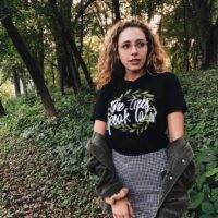 person wearing black The Trees Speak Latin Boyfriend Tee with check black and white skirt and black jean jacket standing outside by trees and ivy