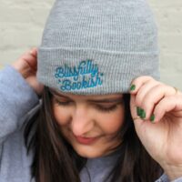 person wearing Blissfully Bookish Beanie standing against white brick wall looking down with hands on their hat