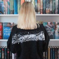 Model in front of a book case wearing a black long sleeve shirt with a white design across the shoulders
