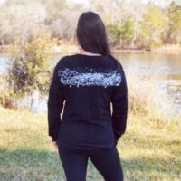 Model in front of a book case wearing a black long sleeve shirt with a white design across the shoulders