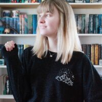 Model in front of a book case wearing a black long sleeve shirt with a white design on left chest