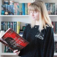 Model in front of a book case wearing a black long sleeve shirt with a white design on left chest