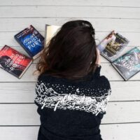 Model in front of a book case wearing a black long sleeve shirt with a white design across the shoulders