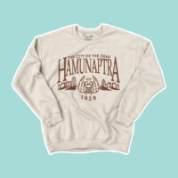 flat picture of a beige sweatshirt with the words "The City of the Dead - Hamunaptra - 1926" with a scarab and Egyptian city in brown ink on a blue background.