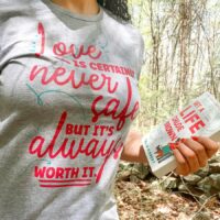 Love is Never Safe grey boyfriend tee with pink text design worn by person holding book in the woods