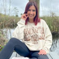 Woman wearing a beige sweatshirt with the words "The City of the Dead - Hamunaptra - 1926" with a scarab and Egyptian city in brown ink.