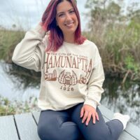 Woman wearing a beige sweatshirt with the words "The City of the Dead - Hamunaptra - 1926" with a scarab and Egyptian city in brown ink.