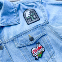 Embroidered patch shaped like a heart with a Welsh dragon with the text "daddy will" on a denim jacket. Embroidered patch shaped with the text "the london institute" and gray building on a denim jacket.